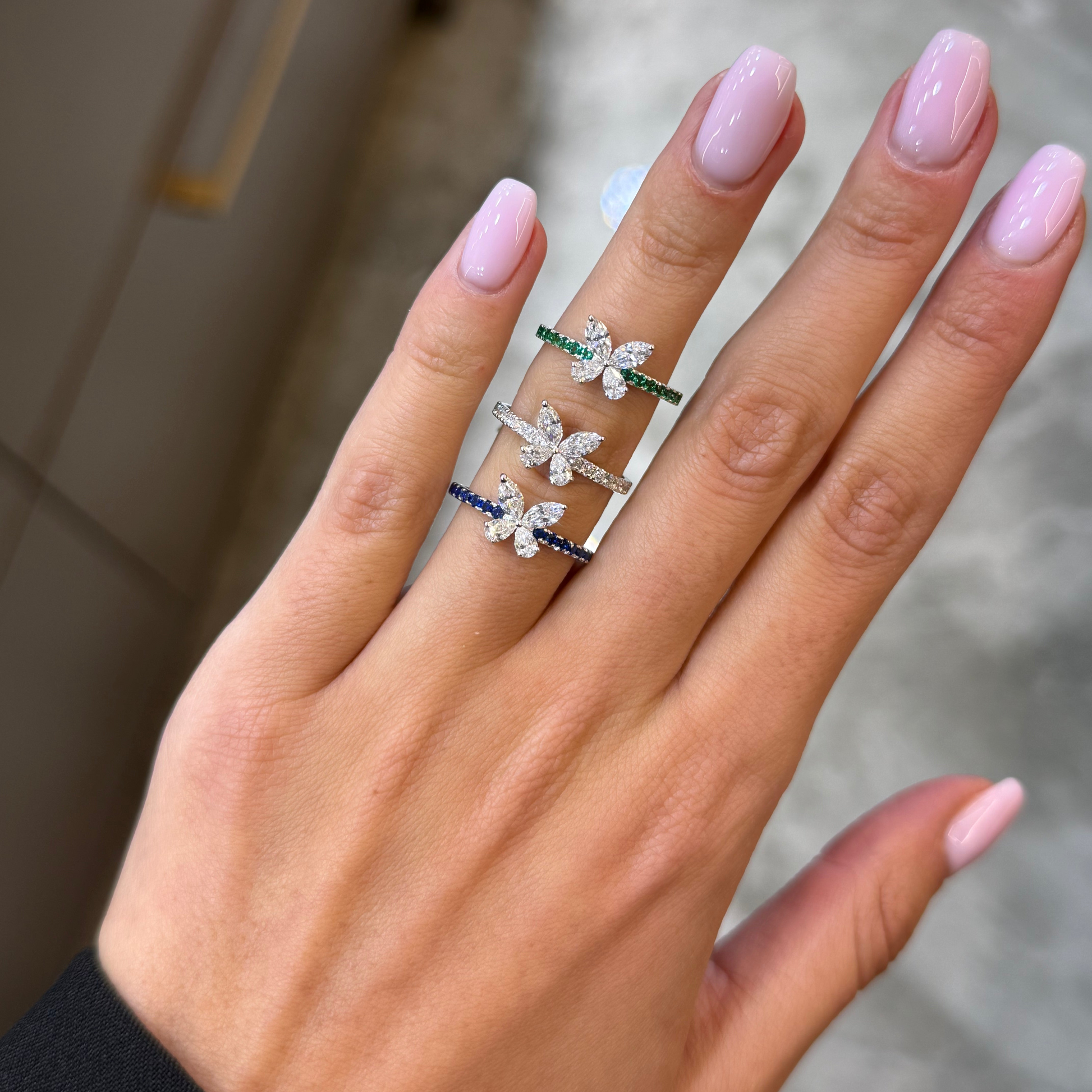 A close-up of a hand with light pink manicured nails wearing three stacked rings. The rings feature silver-toned butterfly or dragonfly motifs with small colored stones: one has green stones, one has blue stones, and one has clear stones.