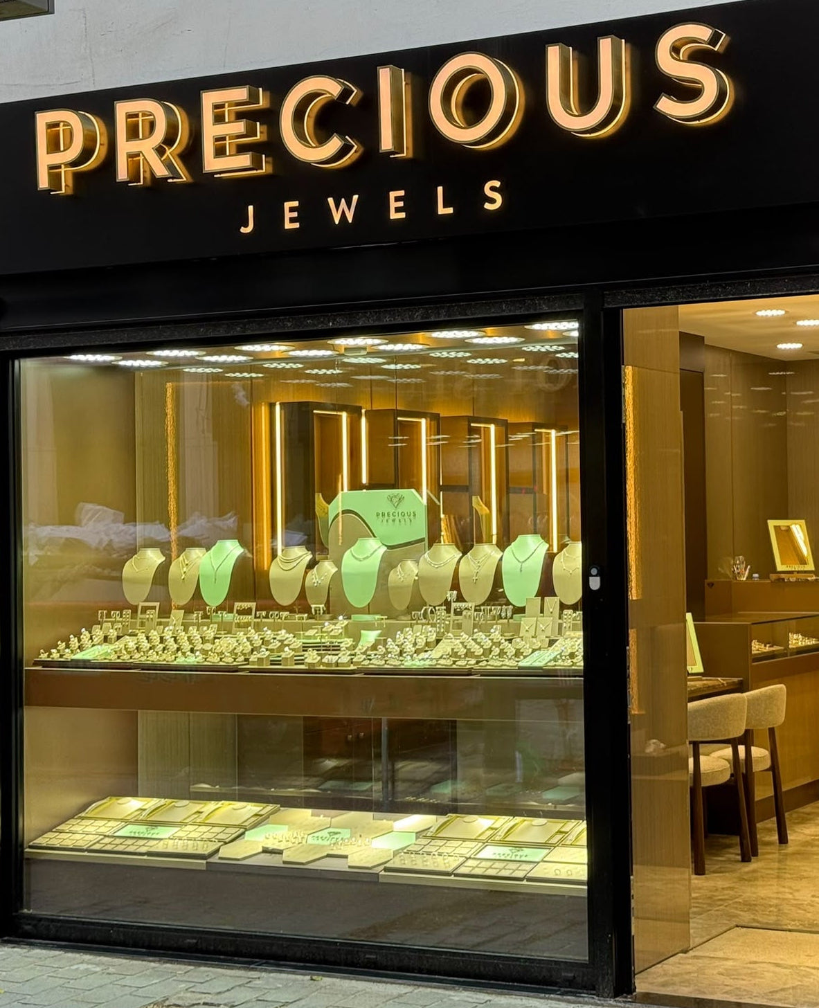 A storefront for Precious Jewels with a black signboard and illuminated gold lettering, large display windows showing jewelry inside, and a gray building facade around it.