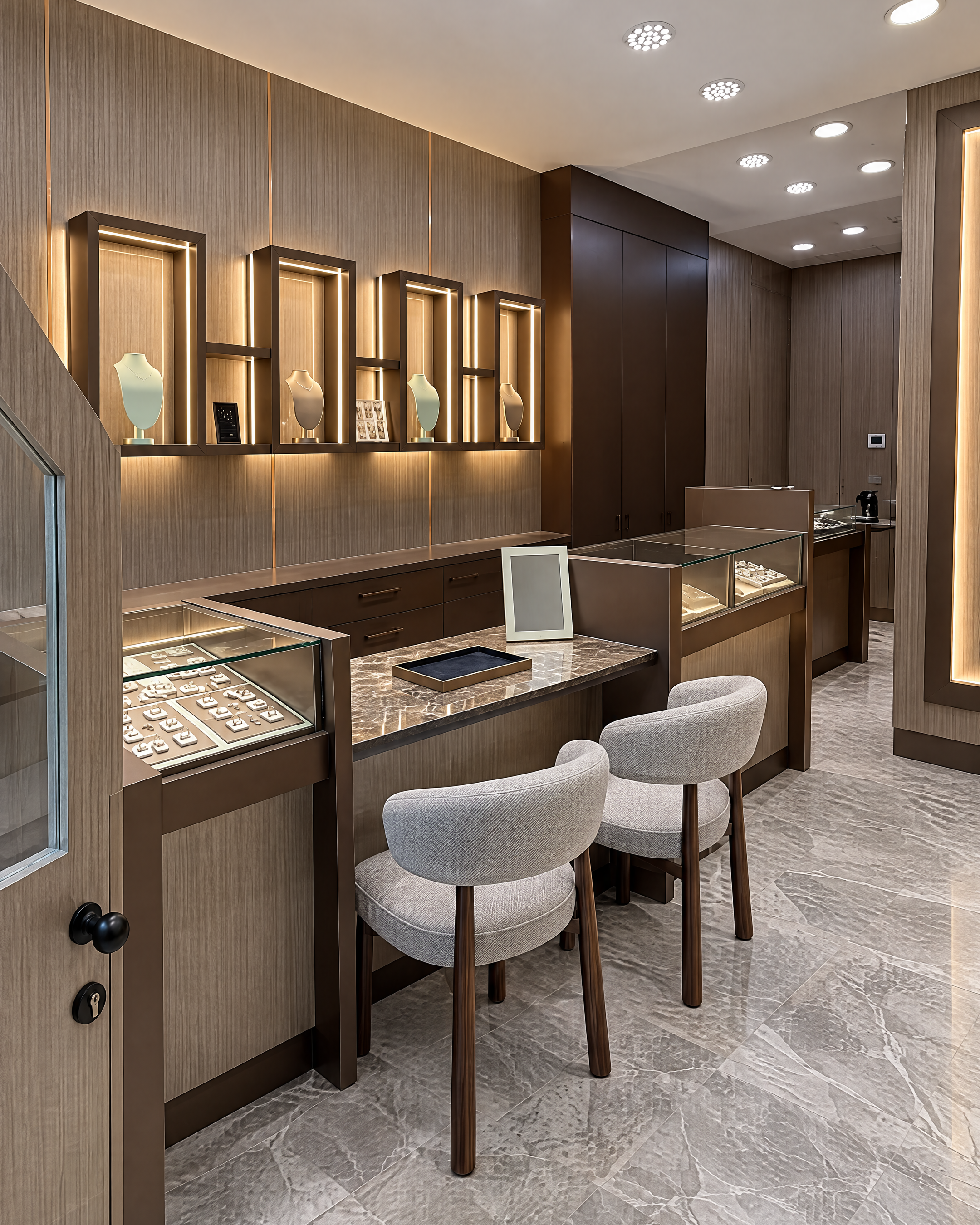 A luxury jewelry boutique interior featuring wood display counters, illuminated glass cases, two upholstered gray chairs, and a polished gray marble floor.