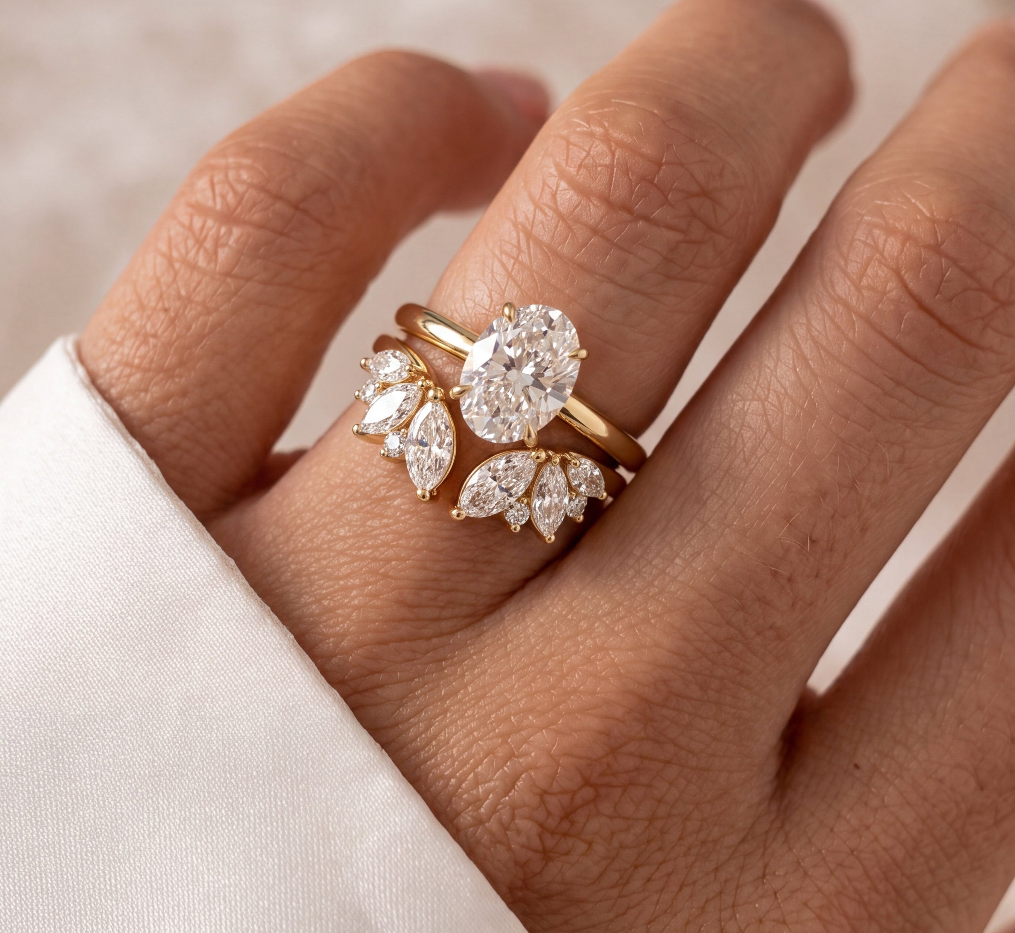 A close-up of a hand wearing a gold ring set with clear gemstones, including a round center stone and marquise-cut side stones, against a soft cream fabric background.